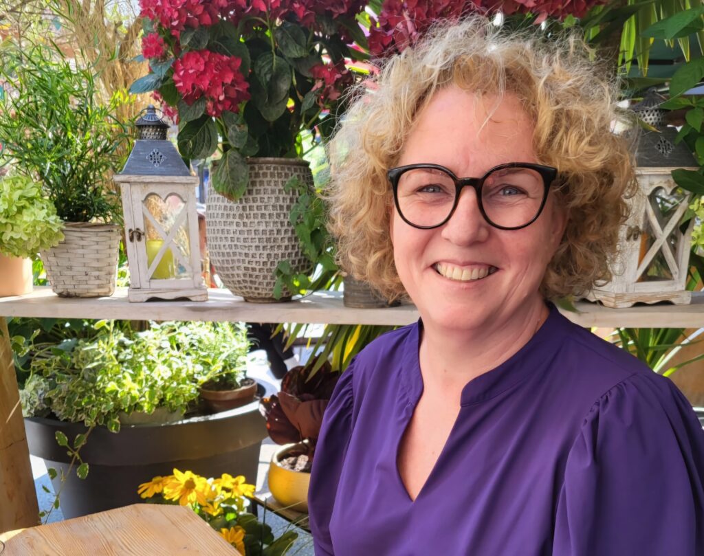 Foto van Jacqueline de Vries met kleurige bloemen in potten op de achtergrond.